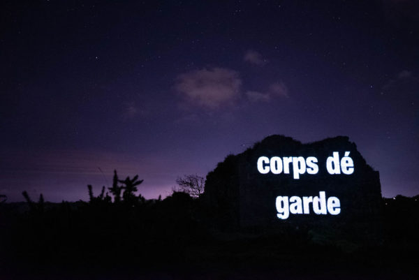 An image of a guardhouse with the Jèrriais word for guardhouse in large letters, projected onto the outdoor wall.